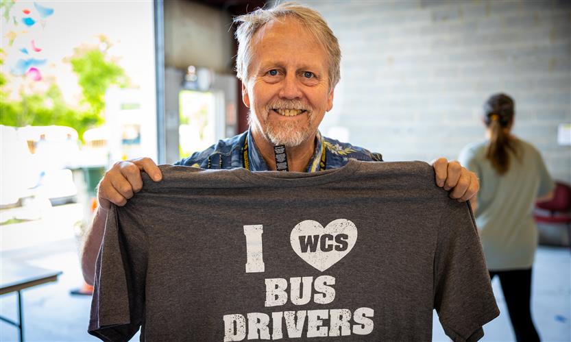 A man smiles while holding a grey shirt that reads \
